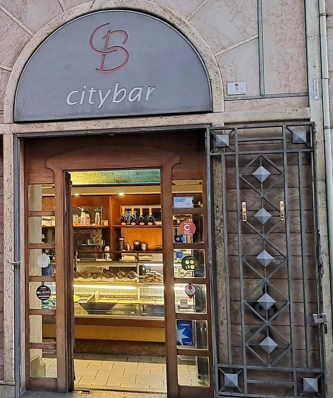 L'ingresso del City Bar in Largo Carlo Felice: l'insegna ad arco color ardesia con il monogramma CB rosso e la scritta citybar, la porta di legno aperta sulla vetrina delle paste e il cancello di ferro a rombi accanto