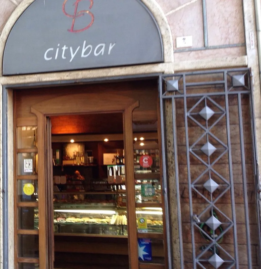 La porta a vetri del City Bar aperta sulla sala: il bancone di legno, la vetrina illuminata dei panini e delle paste